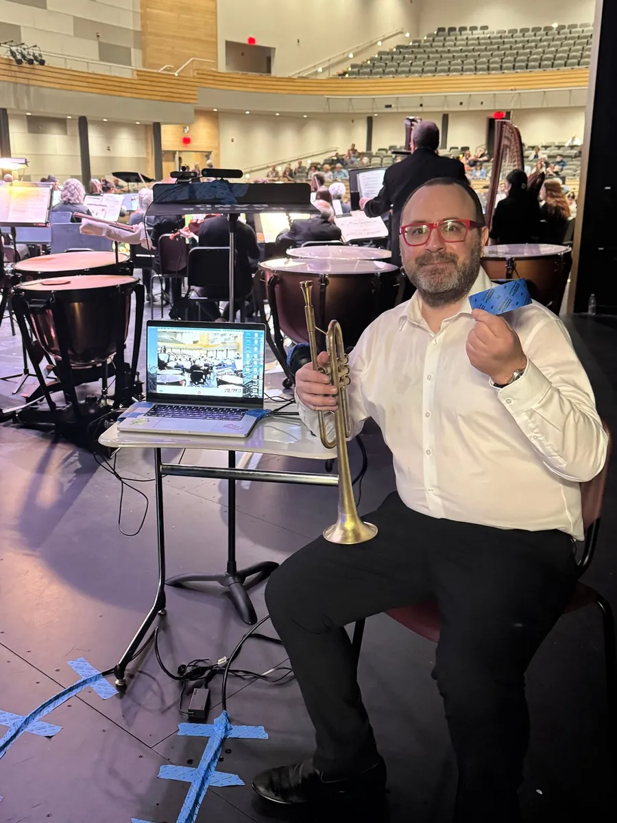 A trumpet player in red glasses and a white shirt sits on stage holding his trumpet and a roll of blue tape, beside a laptop running a video feed on a stand, with timpani, the orchestra, and the audience behind him.