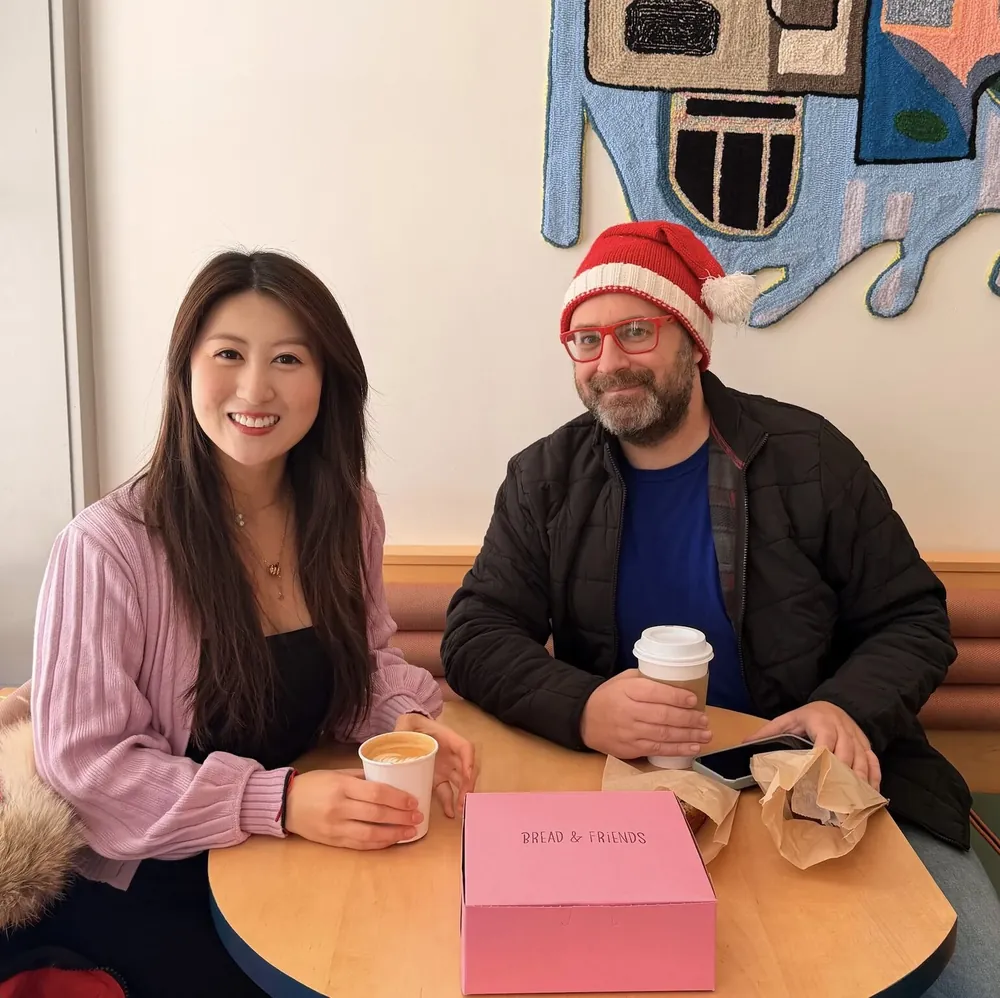 Two people smiling at a round café table with coffee cups and a pink Bread & Friends pastry box; one wears red glasses and a red-and-white knit Santa hat.